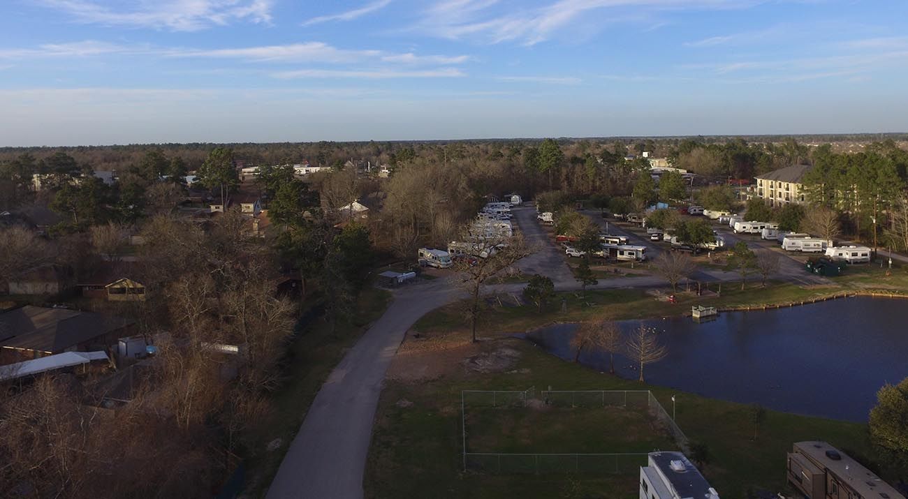Aerial view of a road leading through a residential area with trees, houses, and a lake under a blue sky.