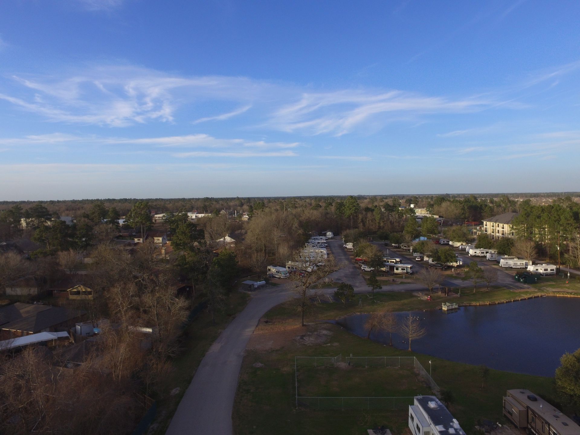 Overhead view of a suburban area with houses, trees, a road, and a lake under a blue sky.