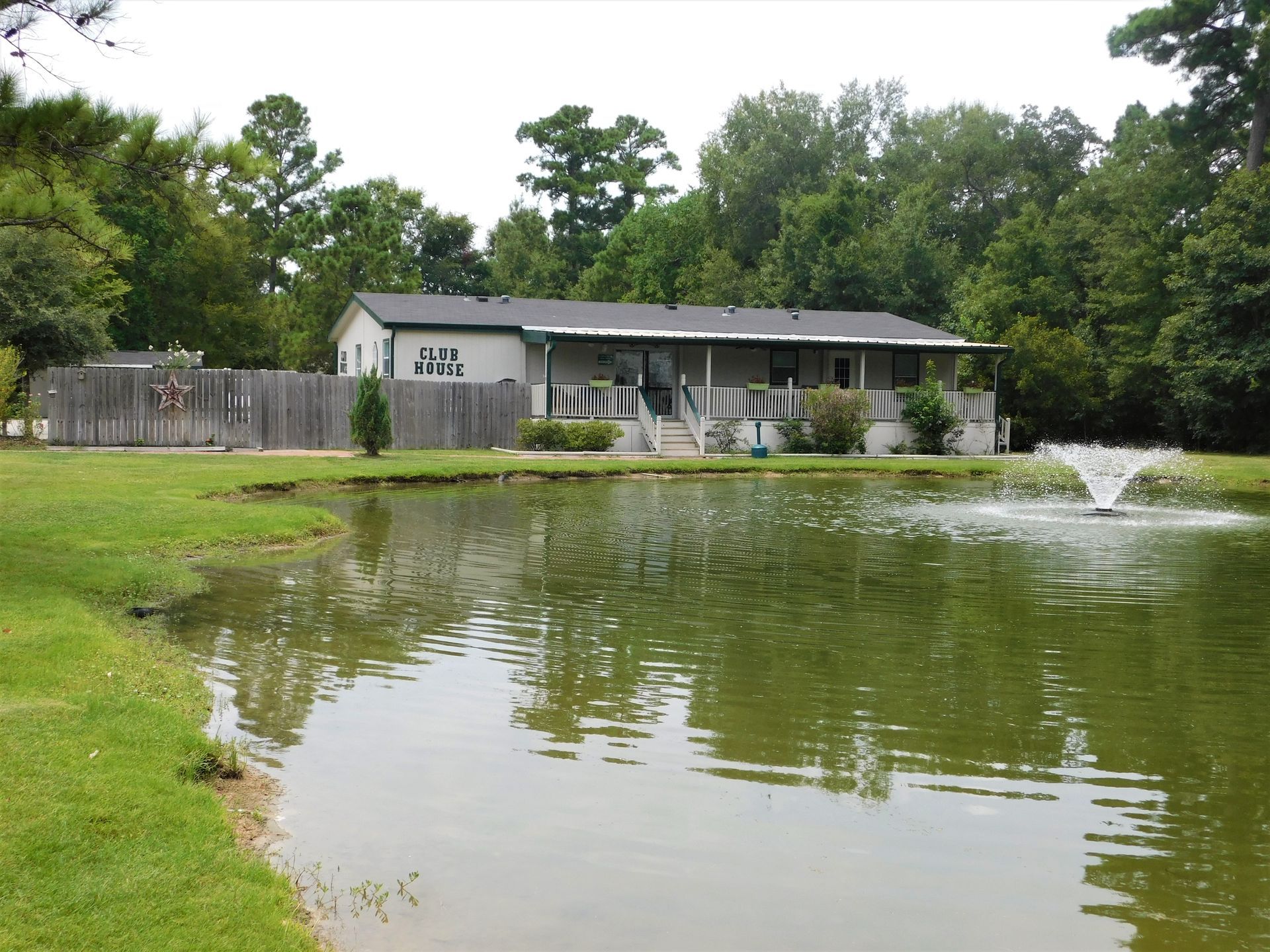 A house with a porch overlooks a pond with a fountain. A wooden fence and trees are visible.