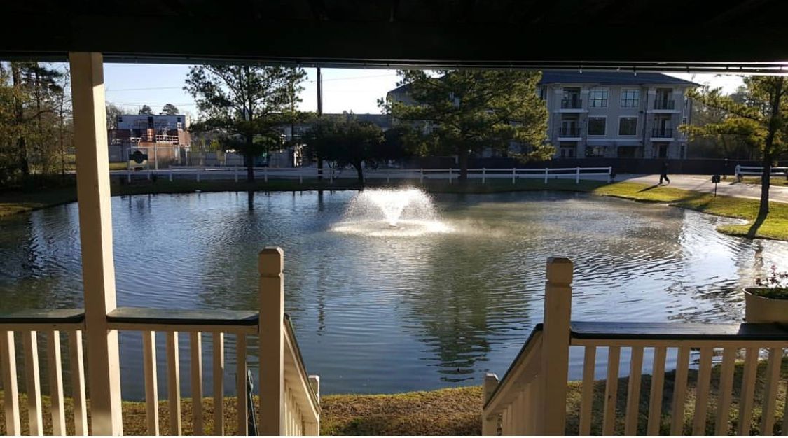 View of a lake with a fountain, seen from a porch. Buildings and trees are in the background.