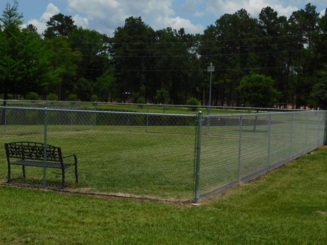 Fenced dog park with bench on grass, surrounded by trees and a cloudy sky.