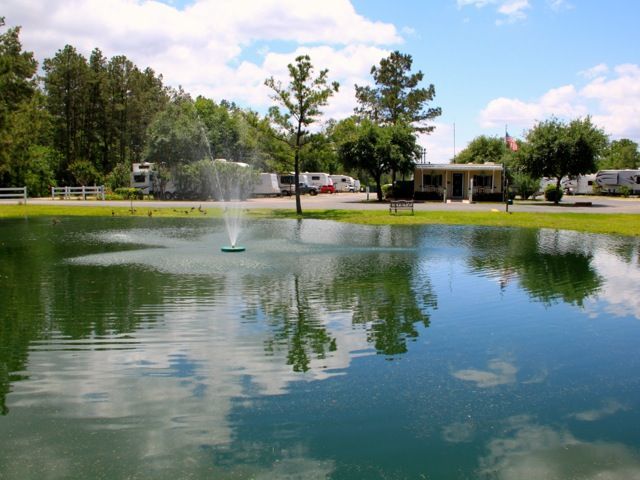 A serene pond with a fountain reflects a campground on a sunny day. RVs and trees are in the background.