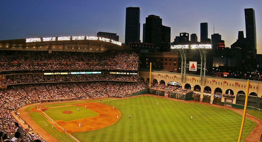 Baseball stadium at dusk, packed with fans, game in progress. City skyline in the background.