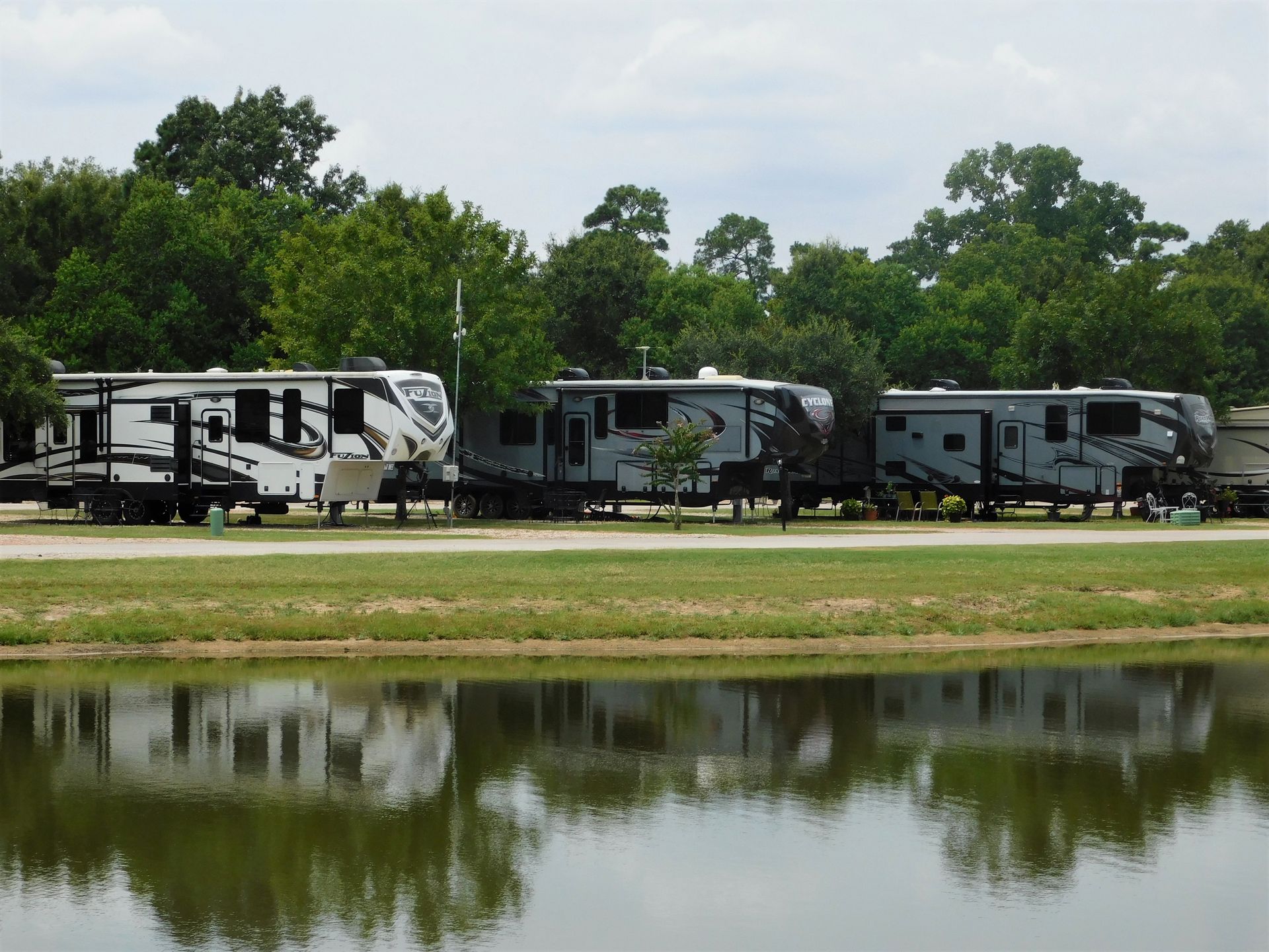 RVs parked lakeside in a campground. Cloudy sky, green trees, water reflecting the vehicles.