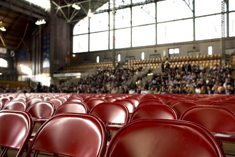 Rows of red folding chairs face a crowd in a large hall with high windows.