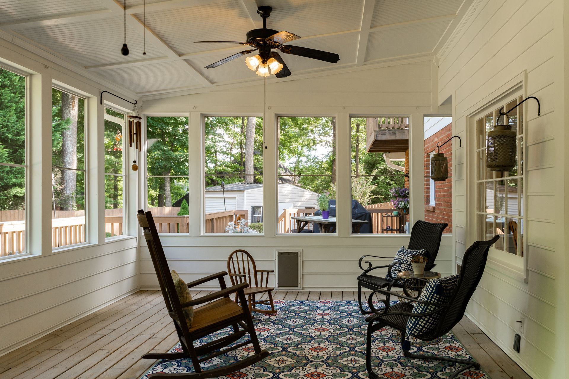 A sunroom with white walls, a ceiling fan, a patterned rug, and several chairs, including a wooden rocking chair. A sunroom with white walls, a ceiling fan, a patterned rug, and several chairs, including a wooden rocking chair.