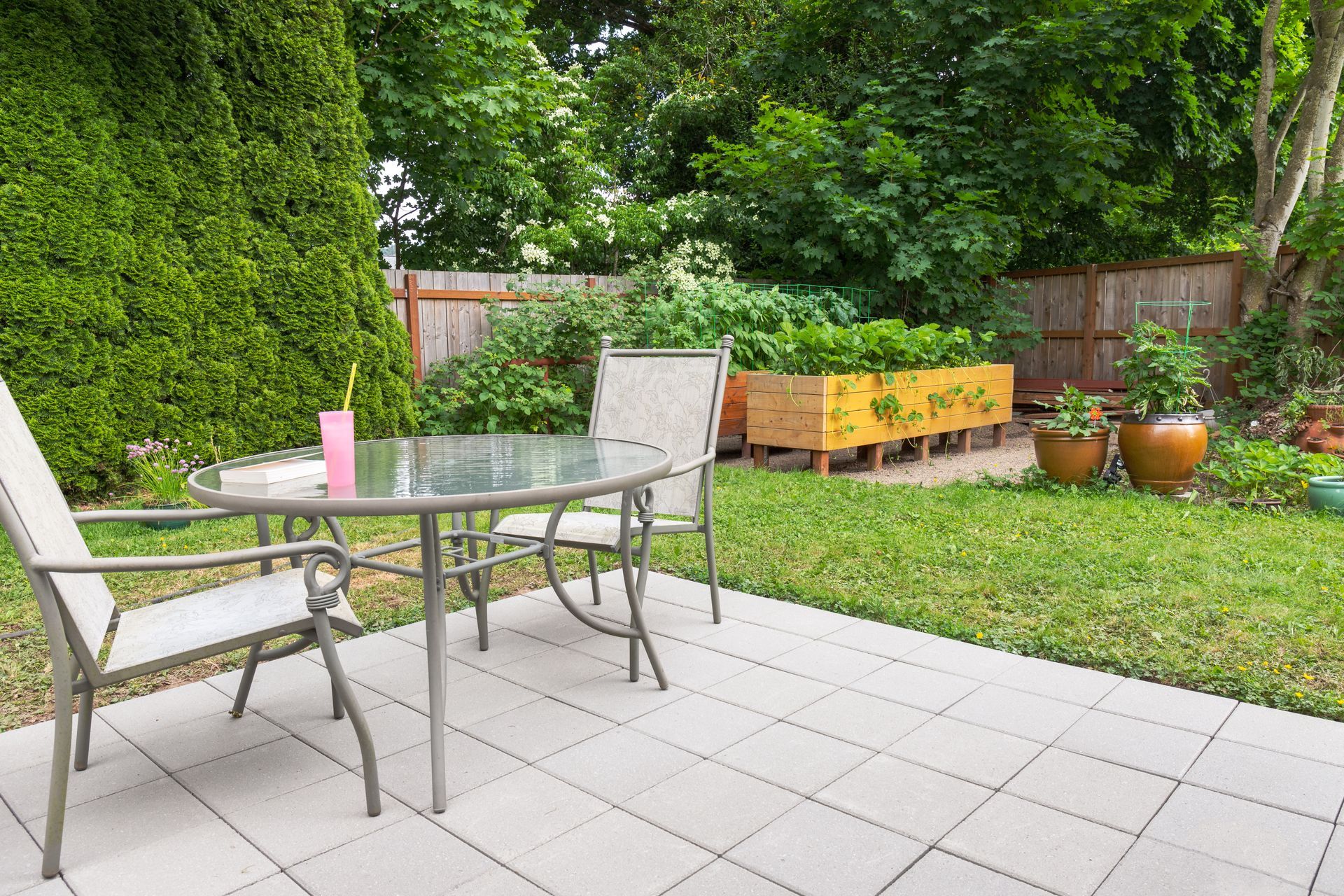 A patio with a round glass table and two chairs sits on a paved area overlooking a green yard with a wooden garden bed.