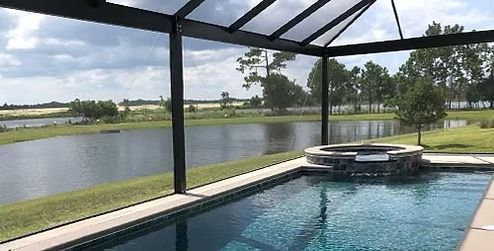 An enclosed pool and hot tub overlooking a scenic lake and green landscape under a cloudy sky.