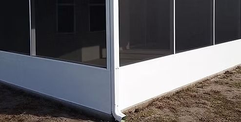 A white aluminum screen enclosure corner with a downspout, positioned on a concrete base over dirt and sparse grass.
