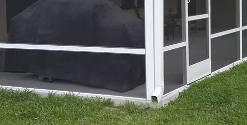 A covered grill sits inside a white-framed screened porch next to a downspout, with green grass in the foreground.