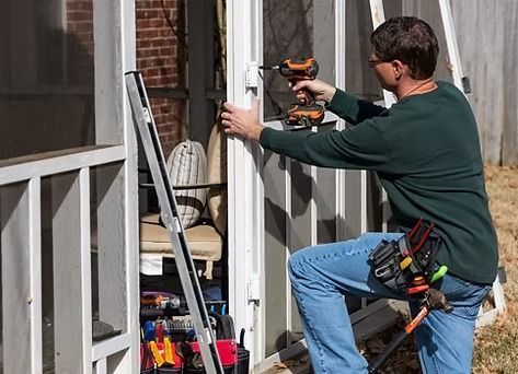 A person in a green long-sleeved shirt uses a power drill to install hardware on a door frame of a screened porch.
