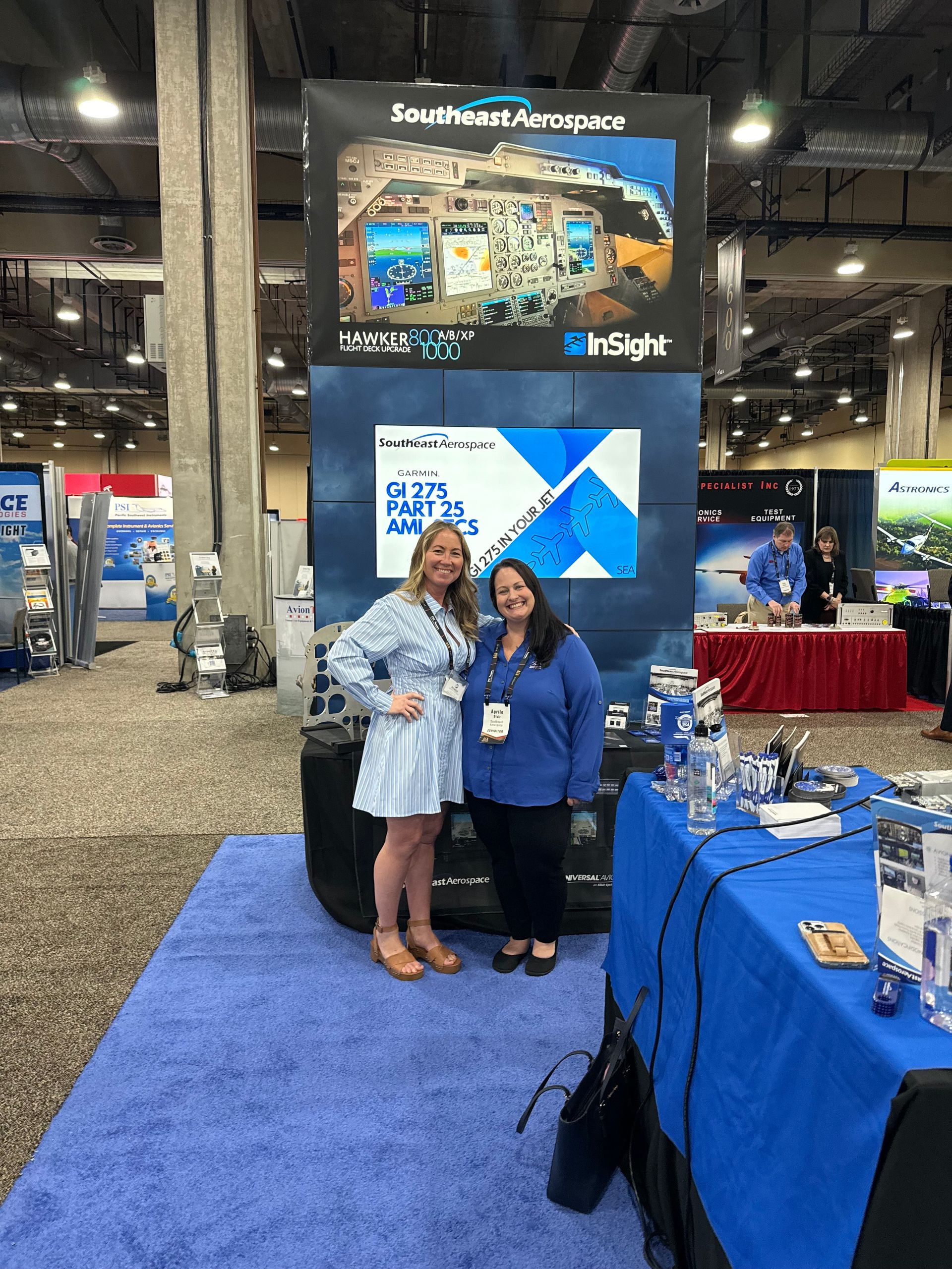 Two women are standing next to each other in front of a sign at a convention.