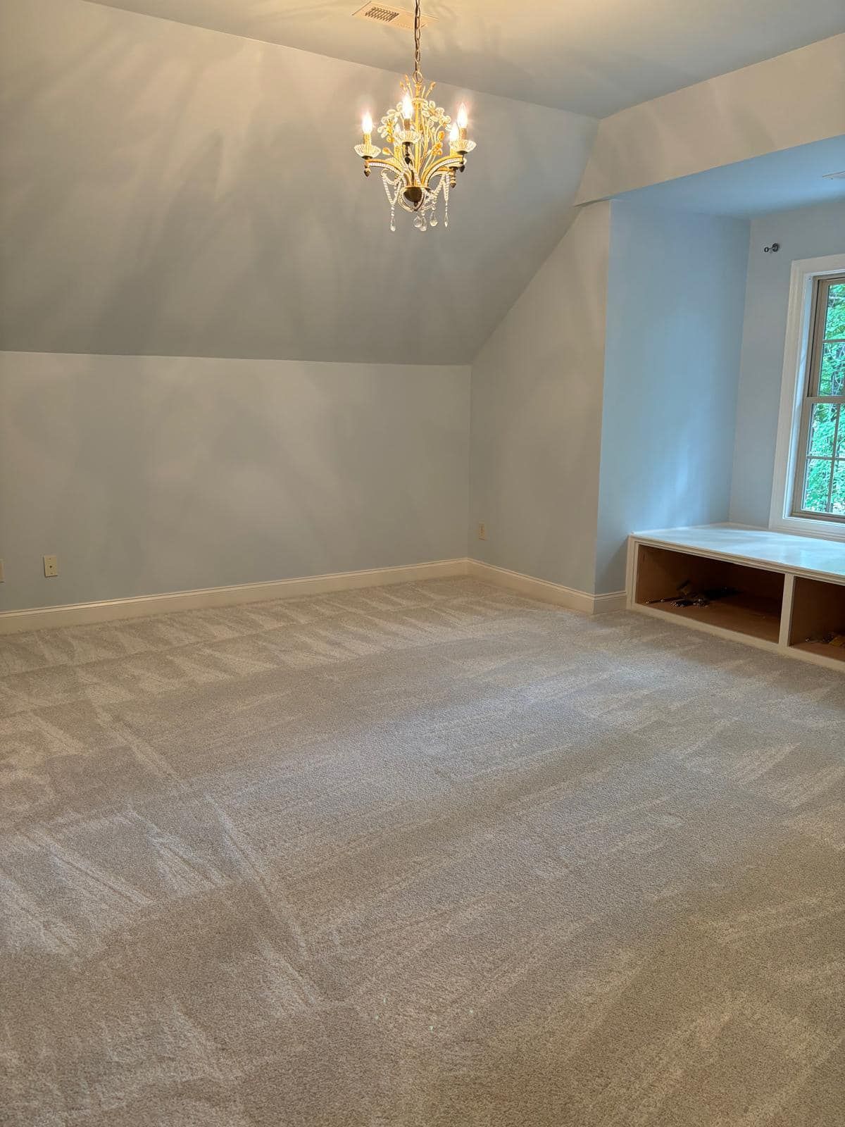 A room with light gray walls and tan carpet, featuring a window seat and a chandelier. The ceiling slopes down.