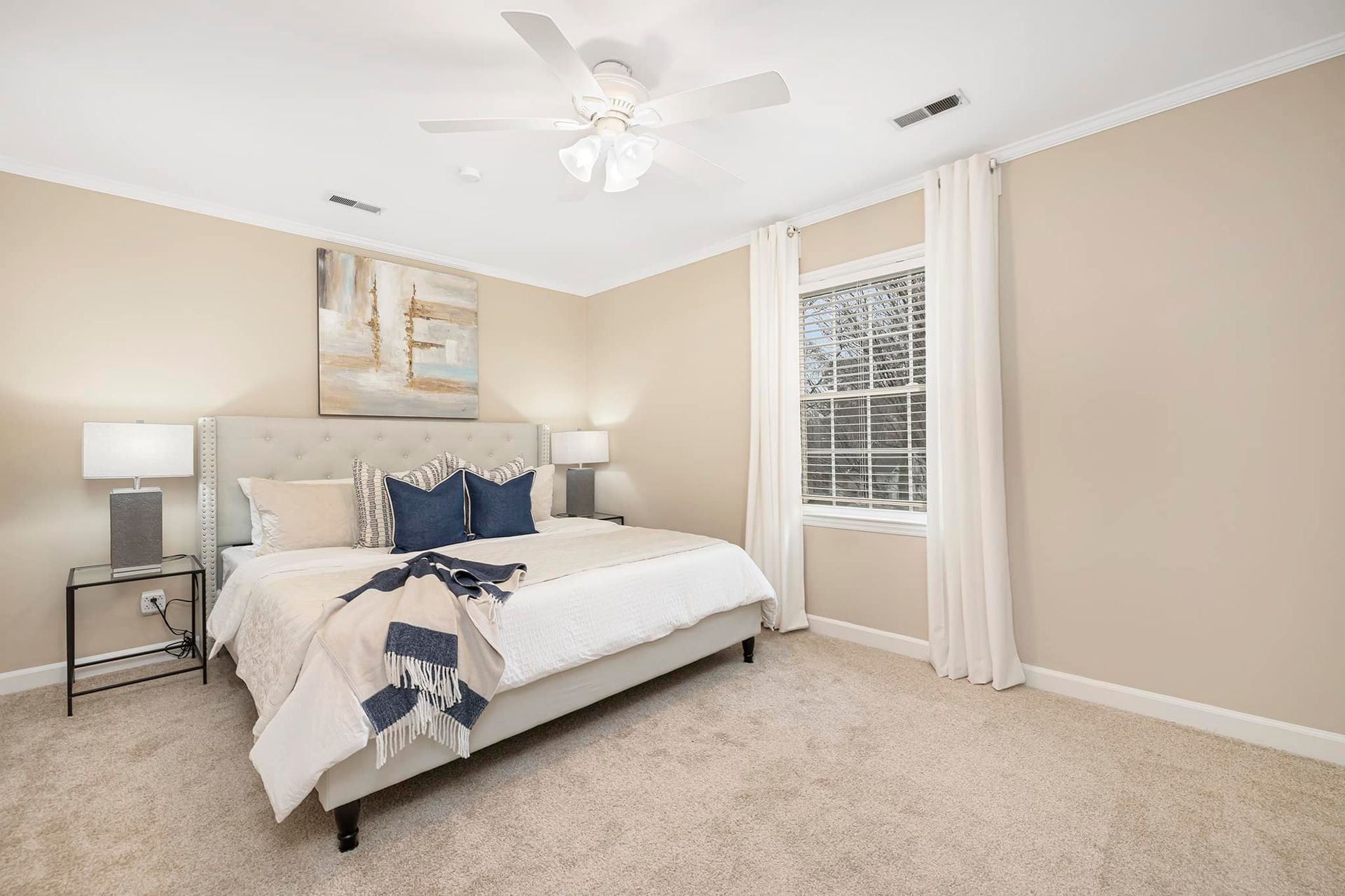 Bedroom with a bed, nightstands, window, and beige walls and carpet. White curtains and a ceiling fan are visible.