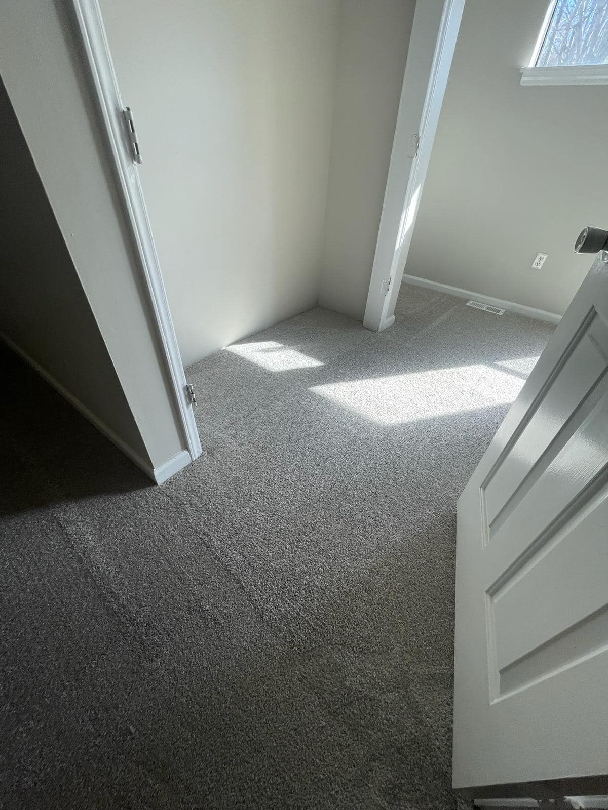 A small room with gray carpet, a white door, and a white support beam. Sunlight streams in from an unseen window.