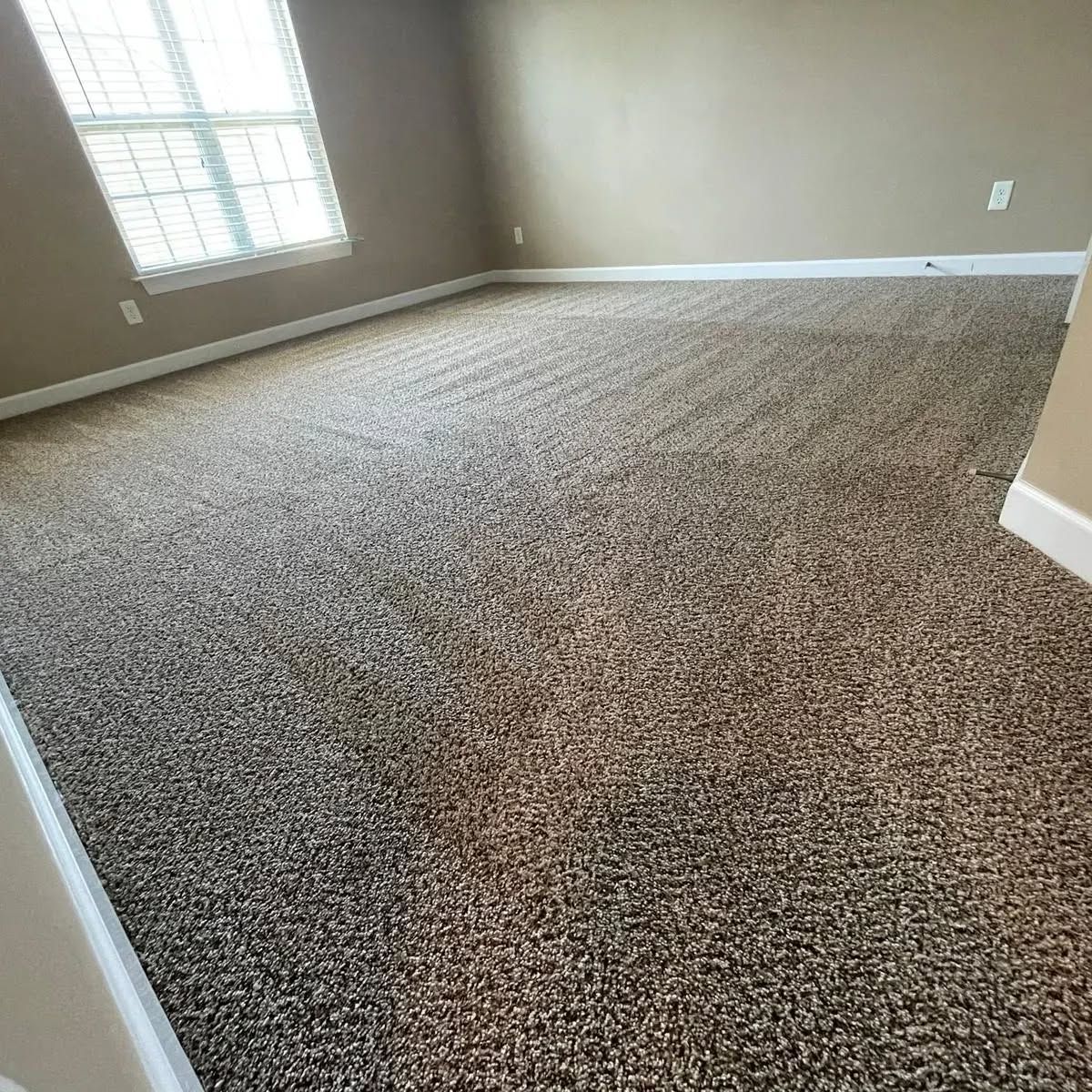 A room with brown and beige speckled carpet, tan walls, and a window with blinds.