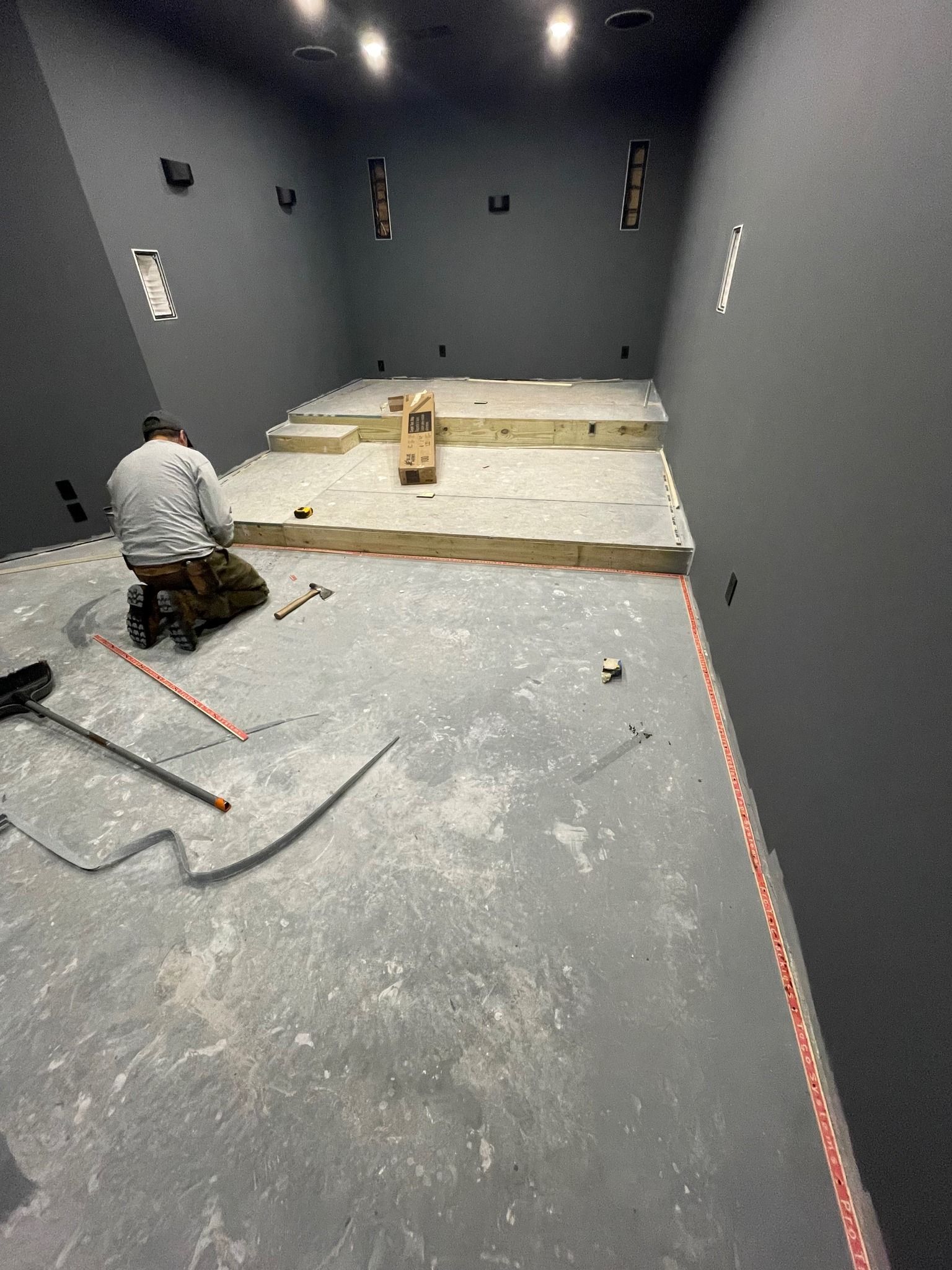 A person kneels on a grey floor inside a dark room. A raised platform is under construction in the center.