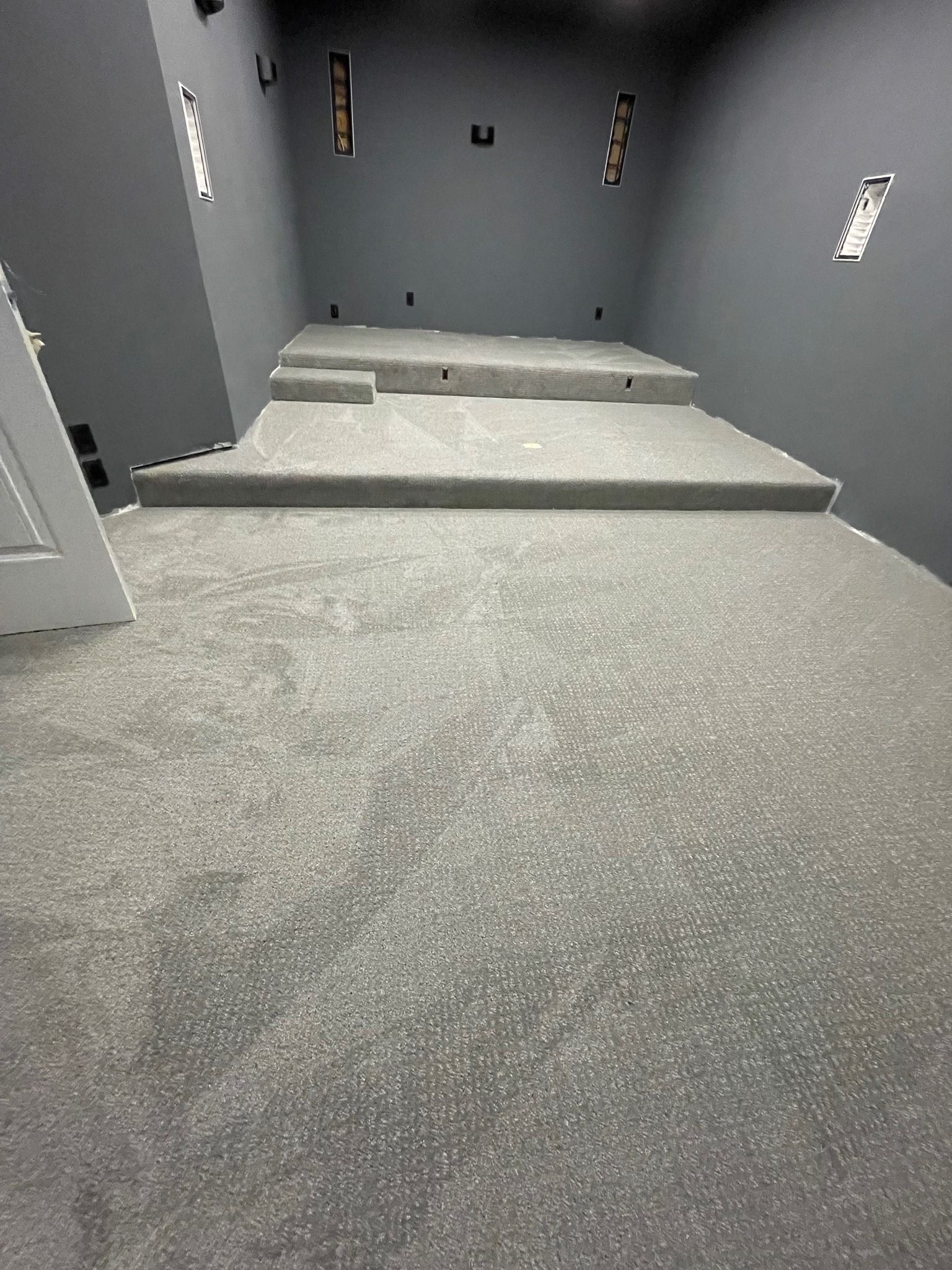 Gray carpeted room with tiered platforms and dark gray walls, possibly a home theater. Empty electrical outlets visible.