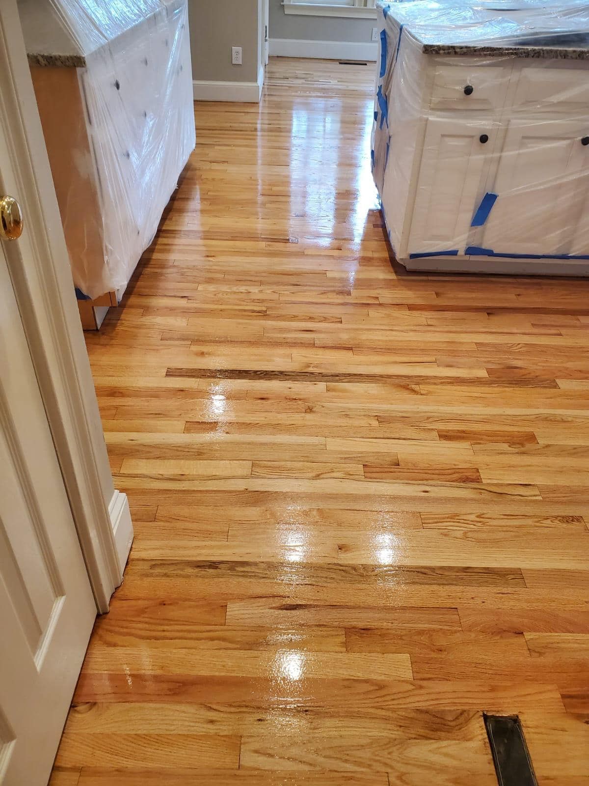 Hardwood floor in a kitchen with white cabinets wrapped in plastic. The floor is a light, glossy wood.