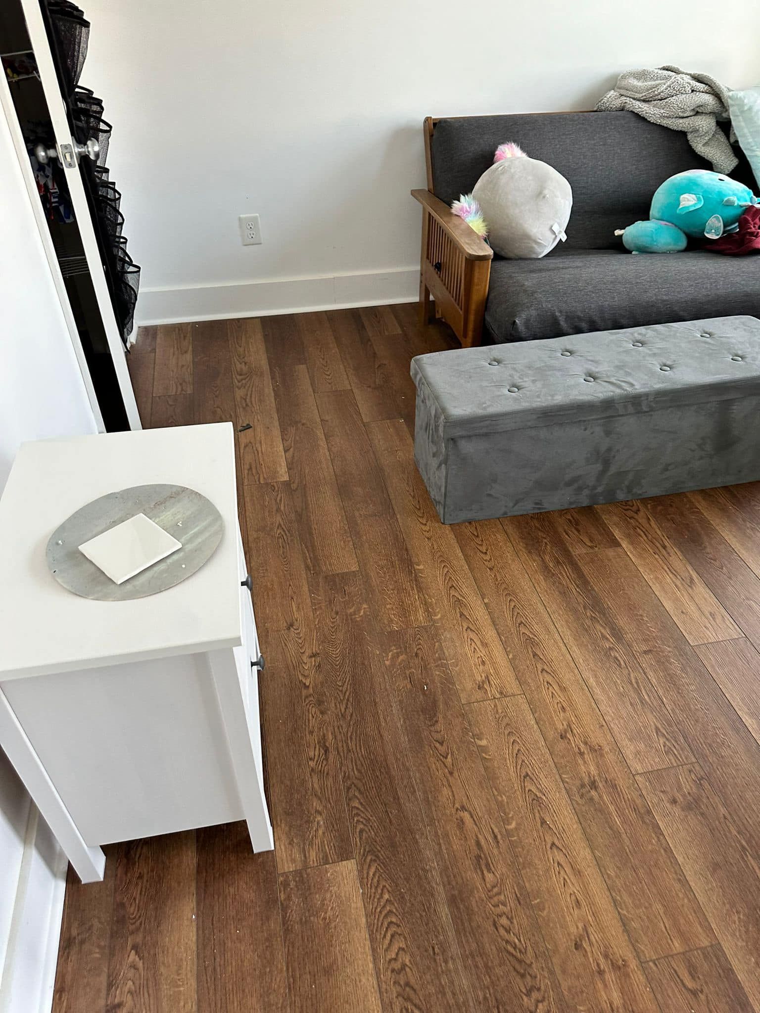 A living room with a futon, gray ottoman, white side table, and wood-look flooring.