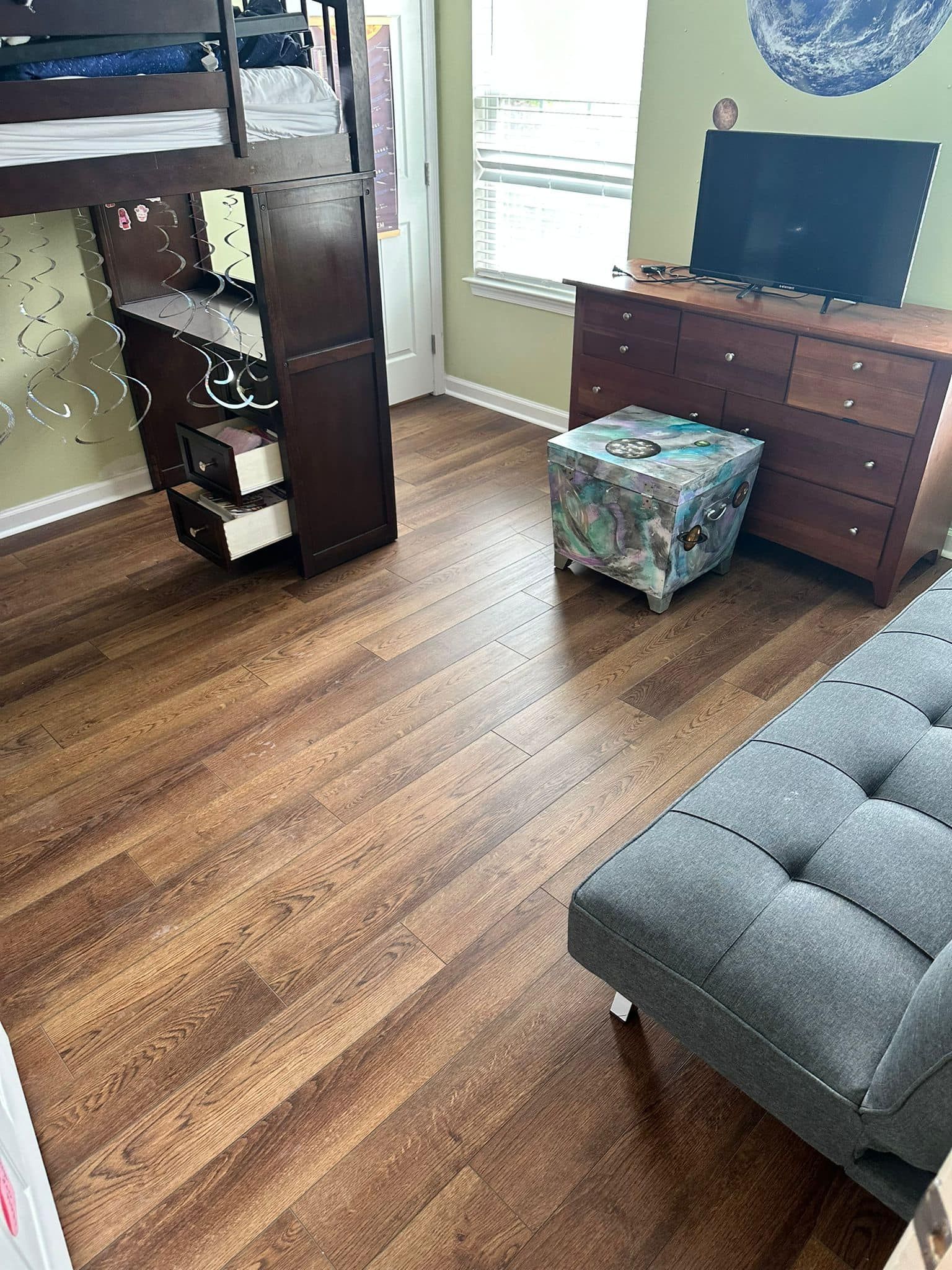 Room with brown wood floor, a loft bed, a dresser, a small cushioned ottoman, and a gray tufted futon.