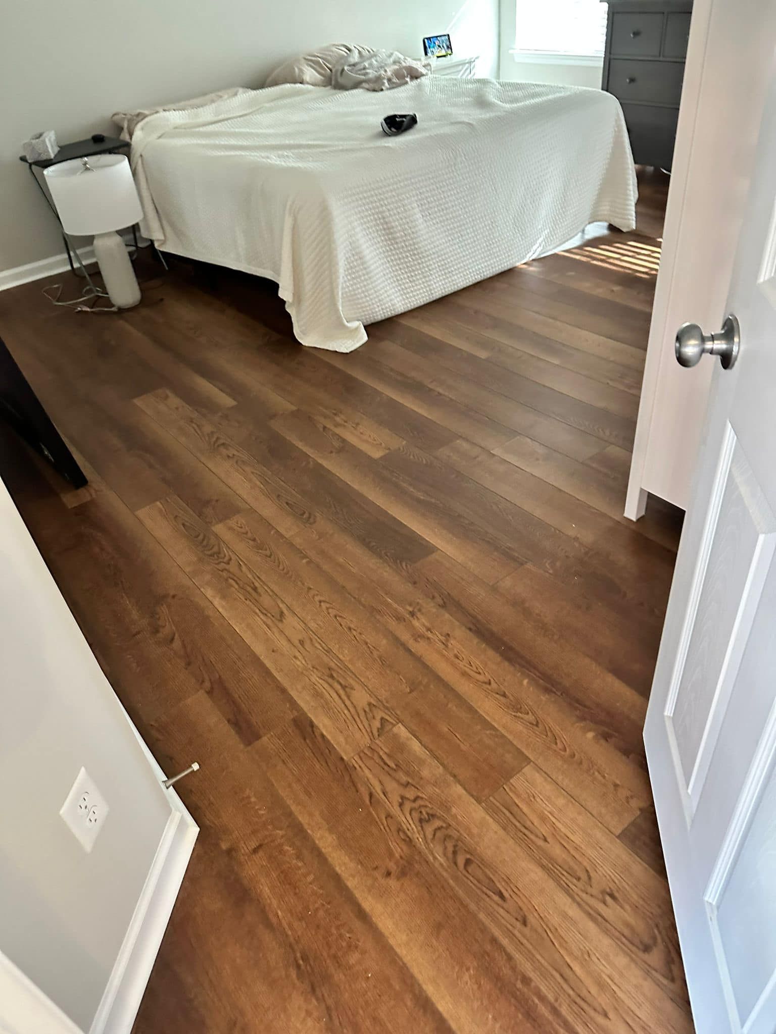 Bedroom with wooden floor. A bed with white sheets sits in the center, with a bedside lamp to the left.