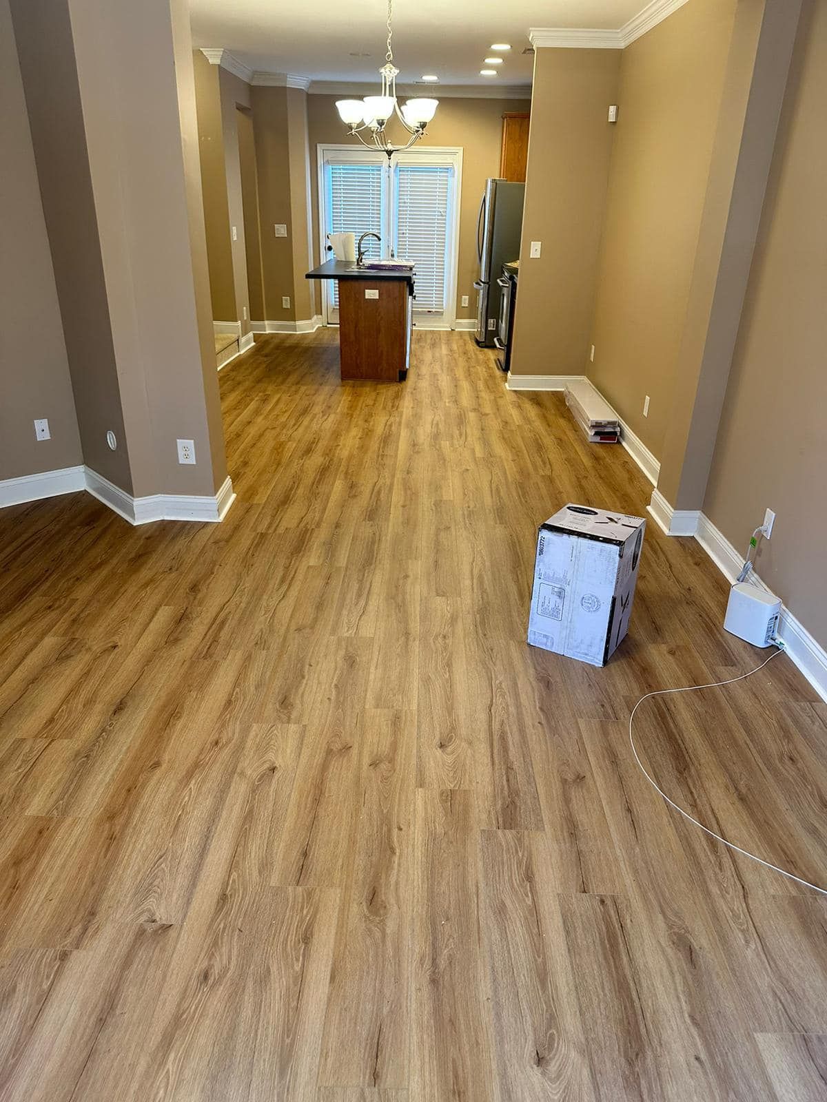 Interior view of a room with light wood-look flooring, beige walls, and a kitchen area visible in the background.