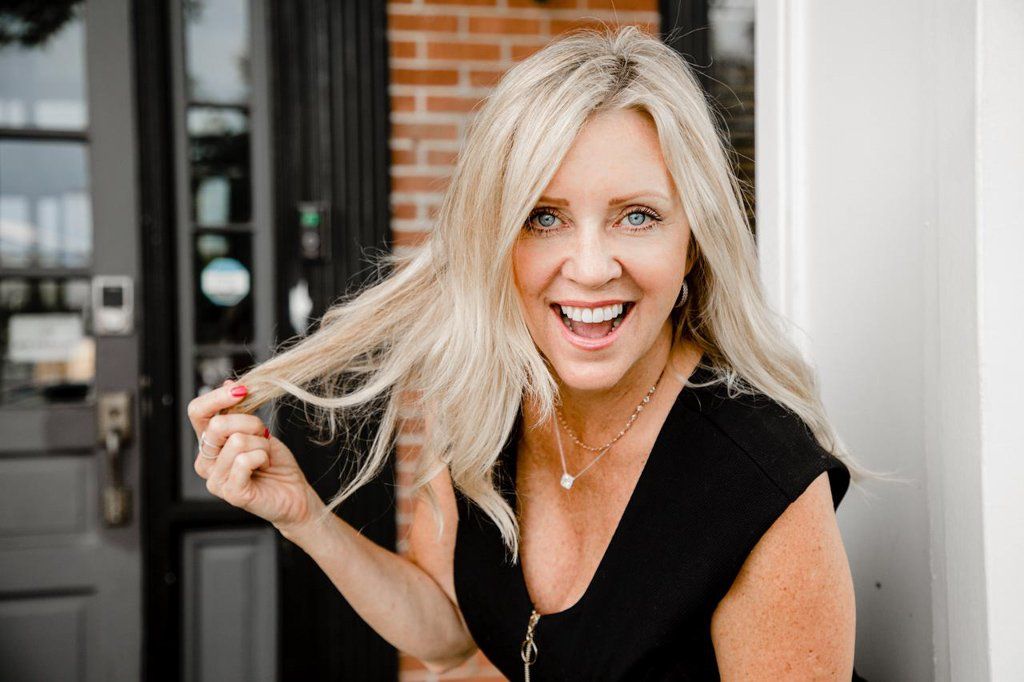 A woman in a black shirt is holding her blonde hair and smiling.
