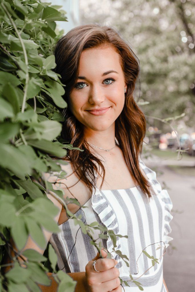 A young woman in a striped dress is standing in front of a bush.