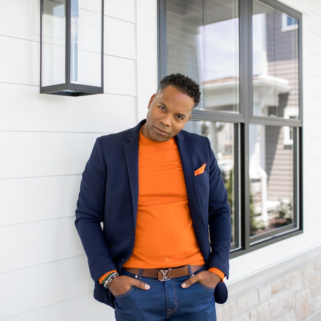 A man wearing an orange sweater and a blue jacket leans against a wall