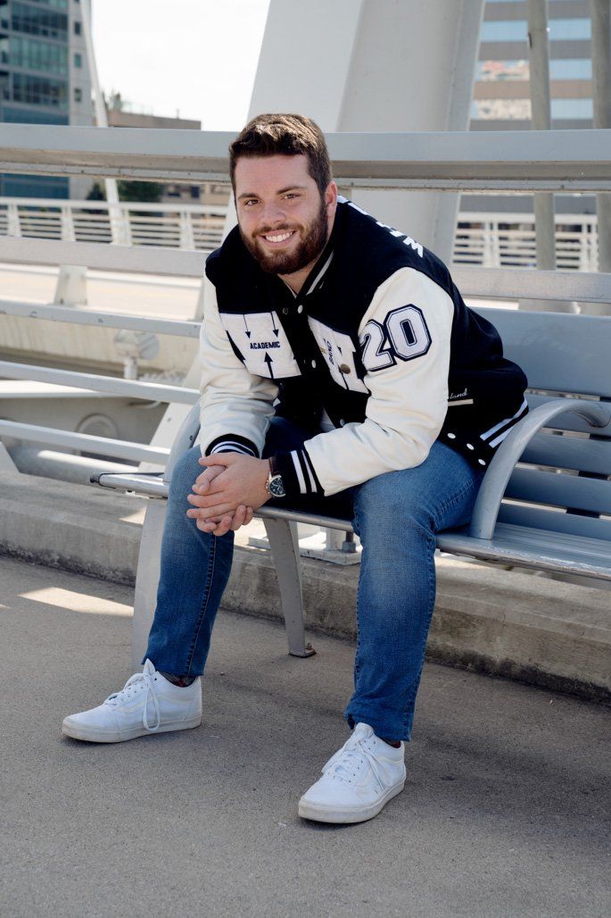 A man wearing a varsity jacket with the number 20 on the sleeves is sitting on a bench