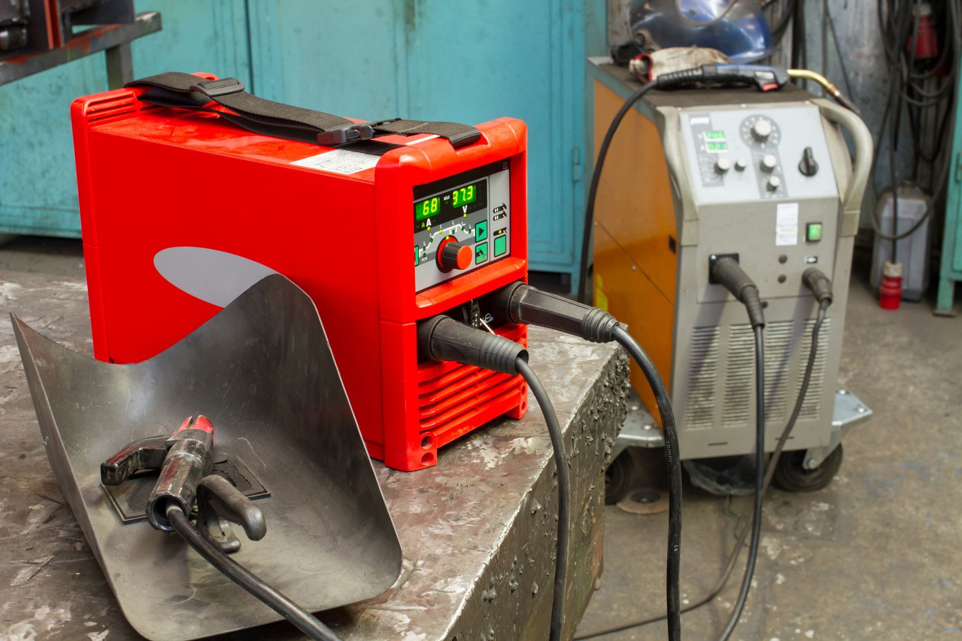 Red and gray welding machines with attached cables on a metal workbench in a workshop