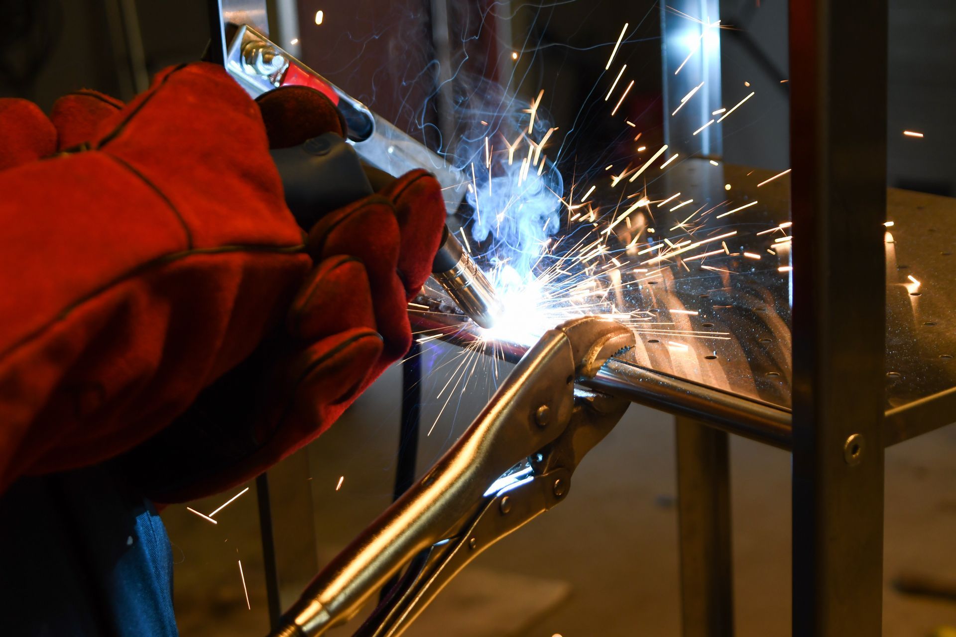 Welder wearing red gloves uses pliers to hold metal pieces while welding, sparks flying