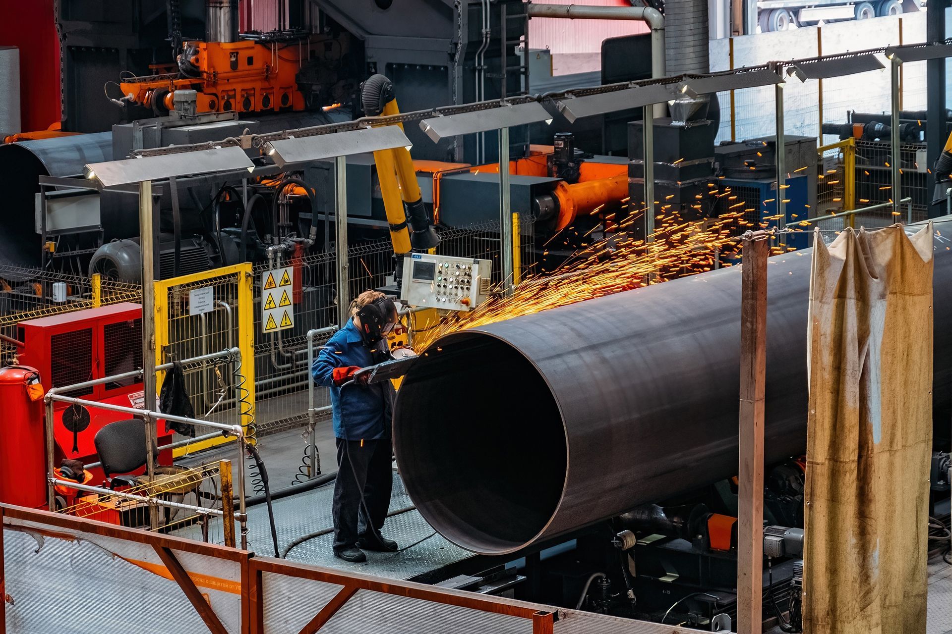 Worker using a grinder on a large metal pipe inside a factory, sparks flying