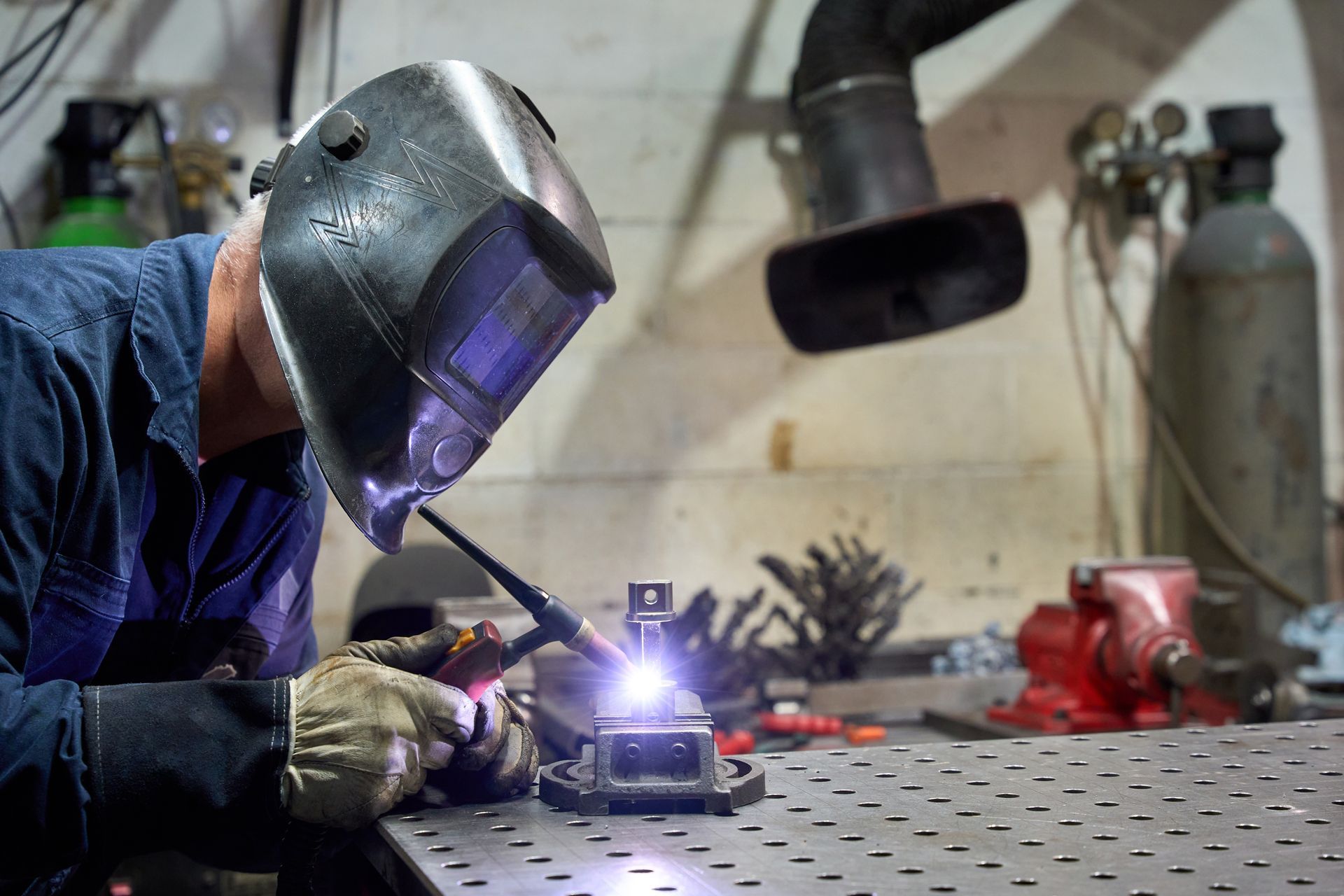 Welder in dark helmet welding metal object on metal table in workshop