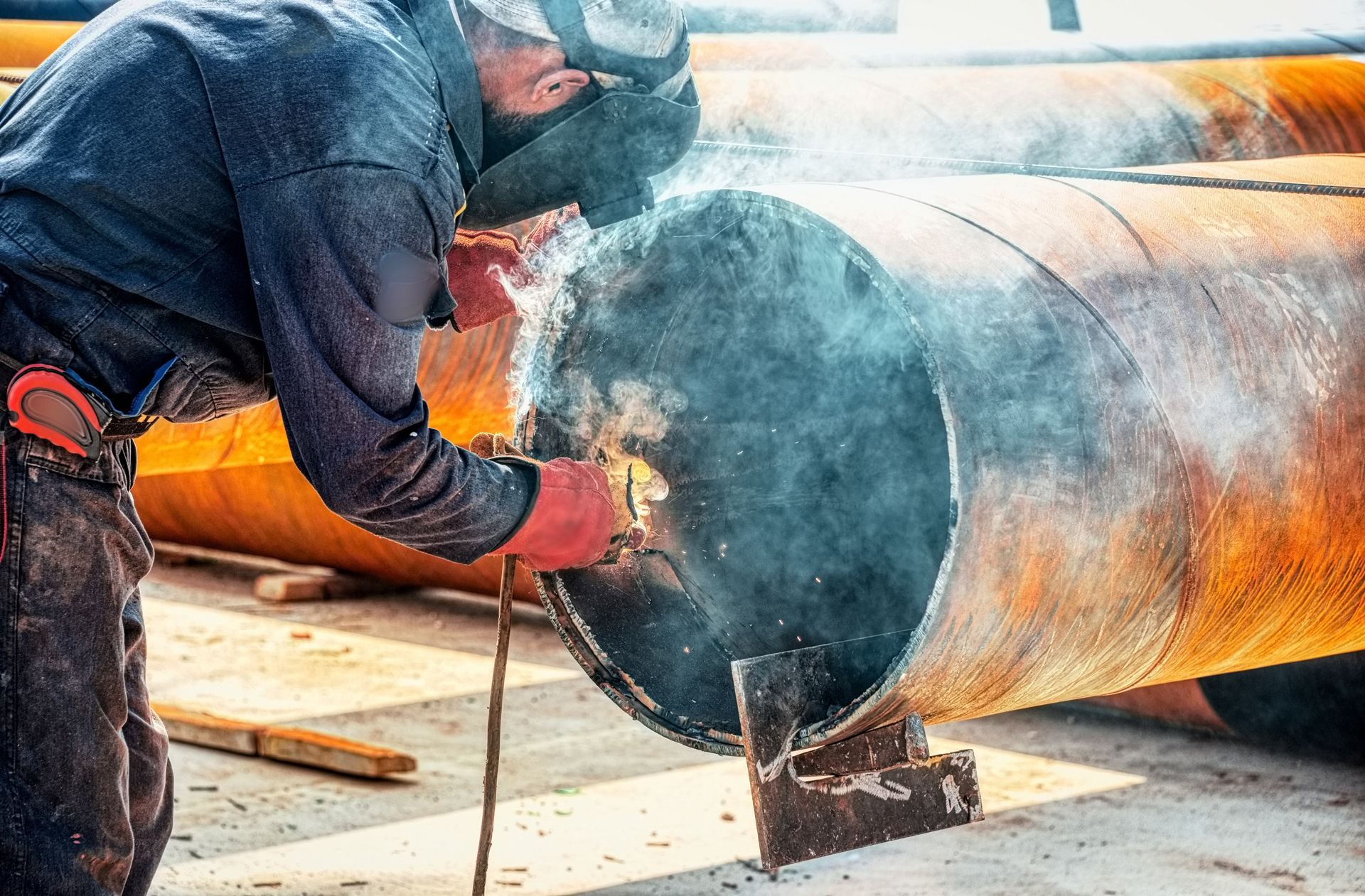 Welder in protective gear using torch to weld a large metal pipe outdoors