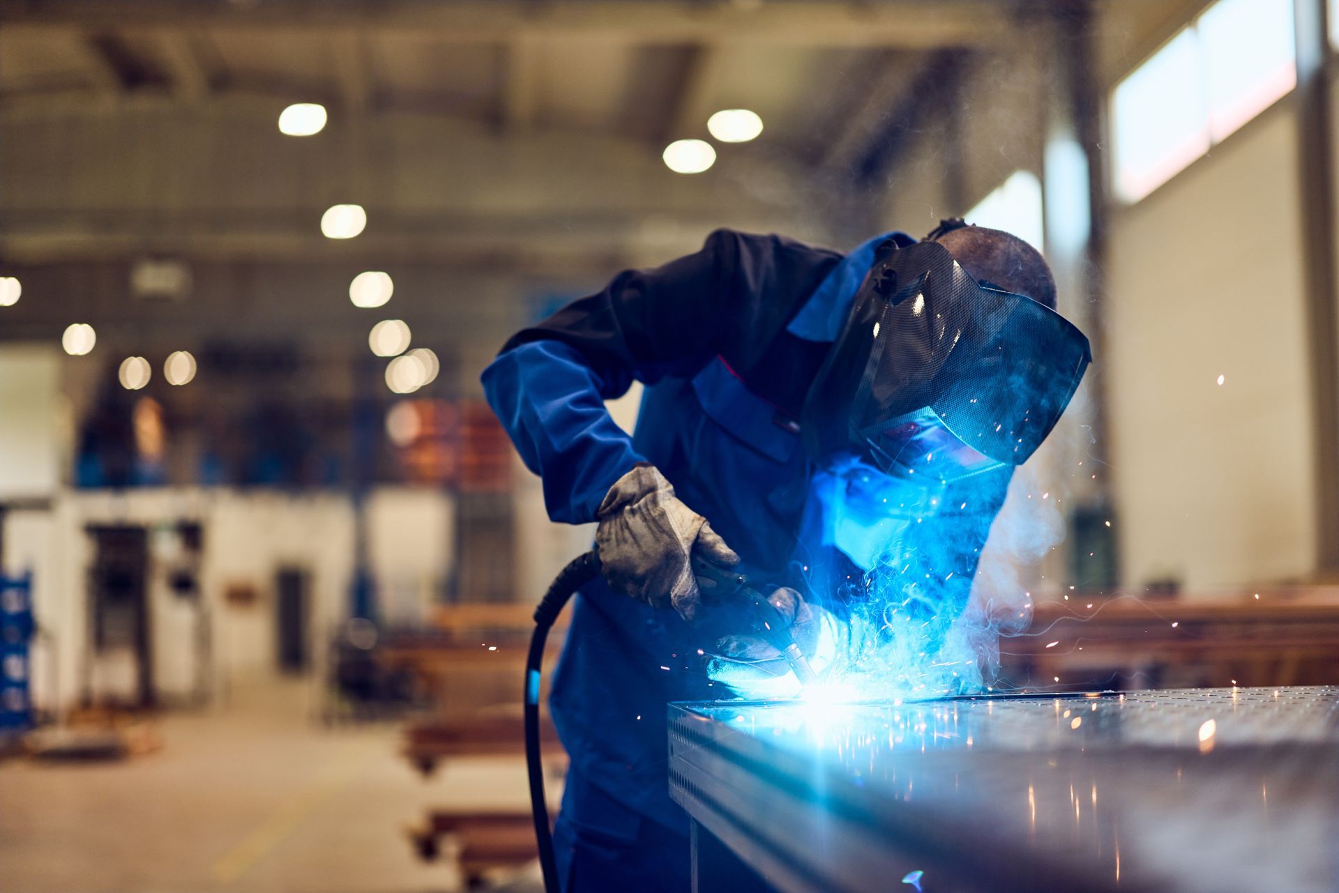 Welder in blue overalls, wearing a mask, welding metal on a table in a workshop, sparks visible
