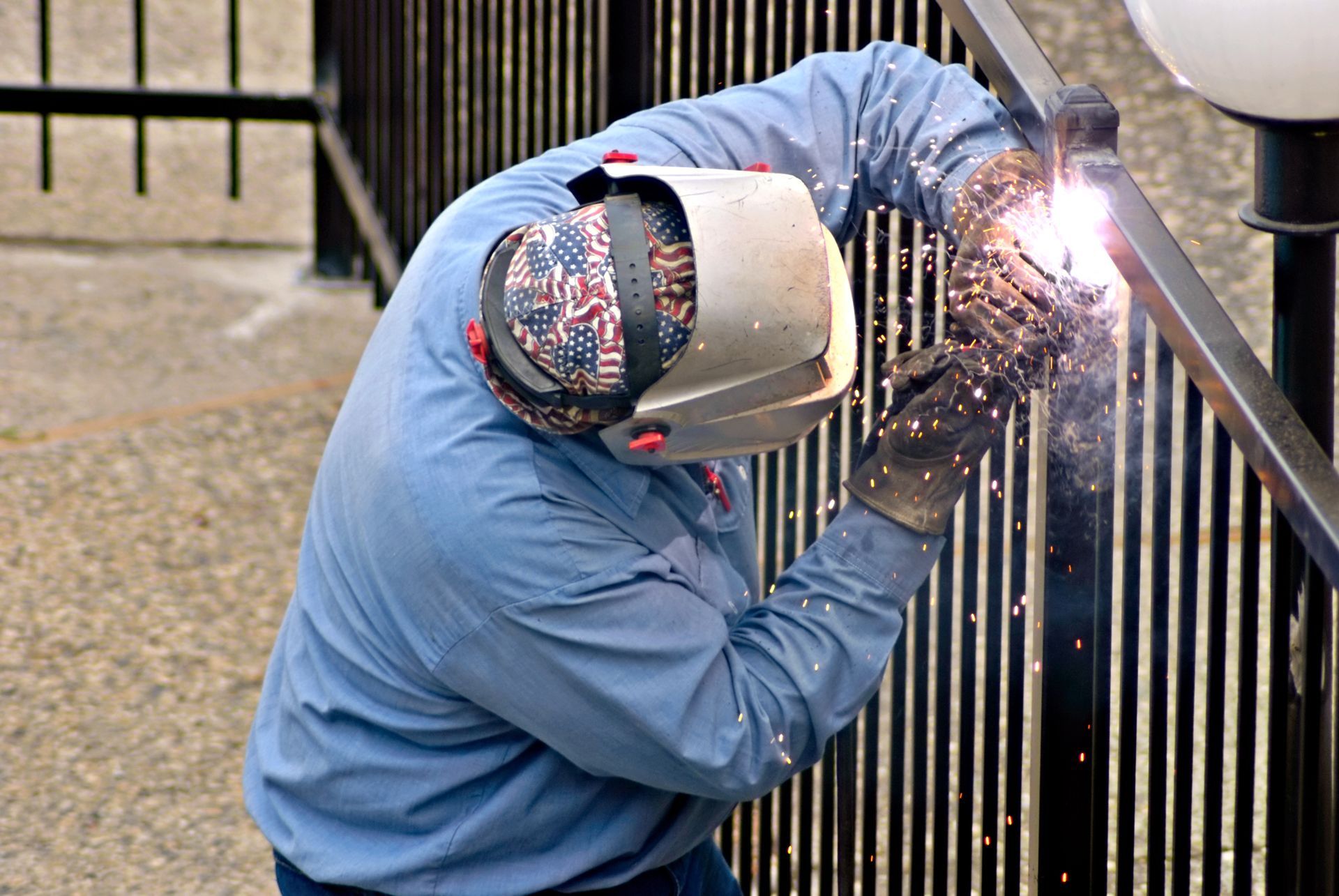 Welder in blue work shirt and protective mask welds a black metal fence