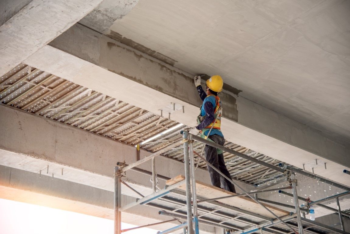 Construction worker on scaffolding, inspecting concrete ceiling, wearing a hard hat and safety harness.