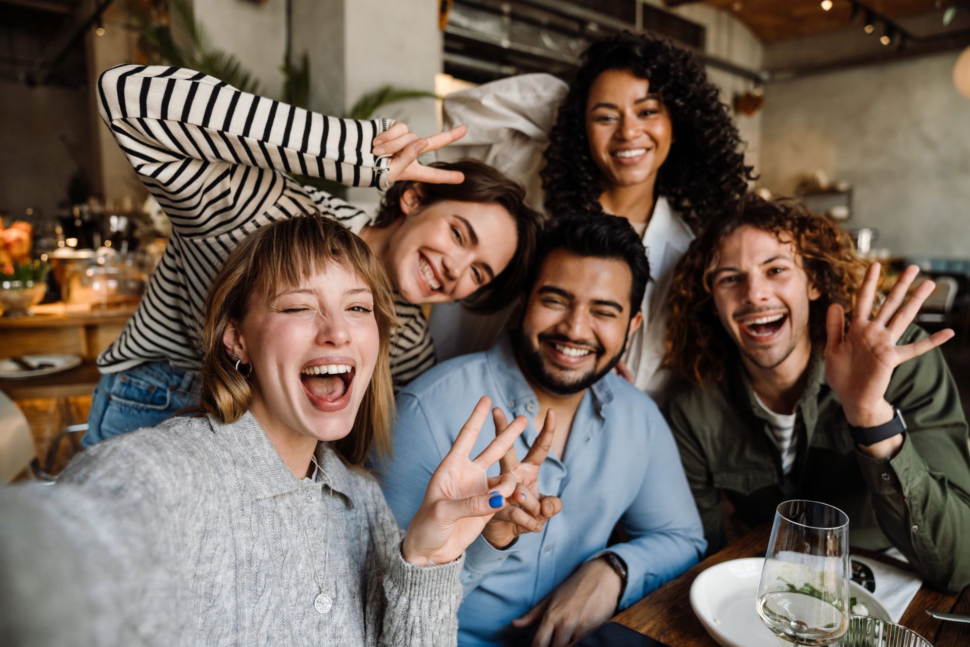 Five smiling people taking a selfie in a restaurant, arms raised, making peace signs.
