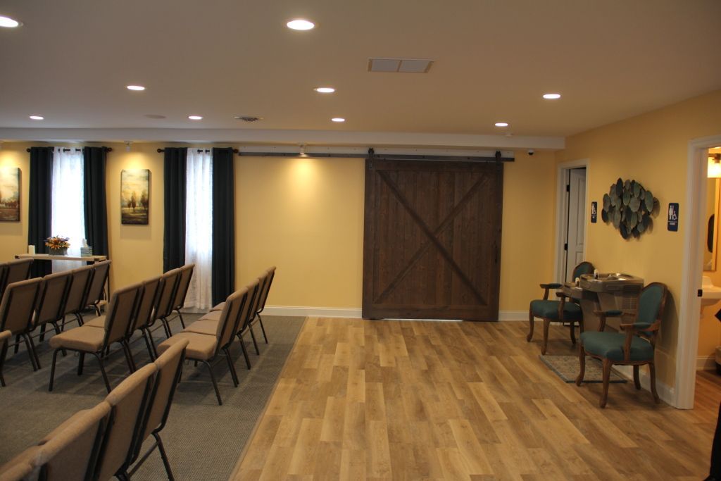 An interior room with a light wood floor, yellow walls, and a dark brown barn door. Rows of chairs are arranged in front of a window with black curtains.