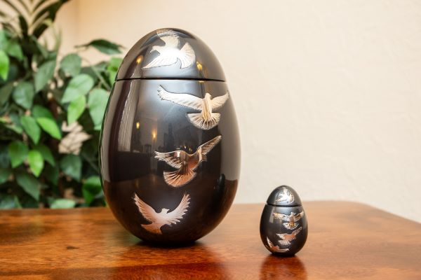Two black egg-shaped urns, one large and one small, decorated with silver flying doves, sit on a wooden table.