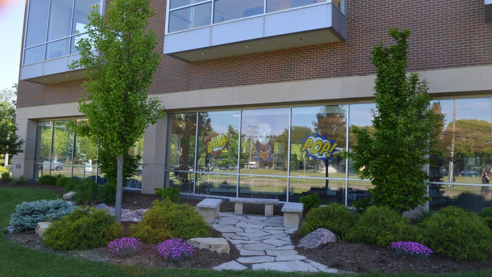 A modern building with large windows reflects trees and a park; a stone path leads to a bench in the garden.