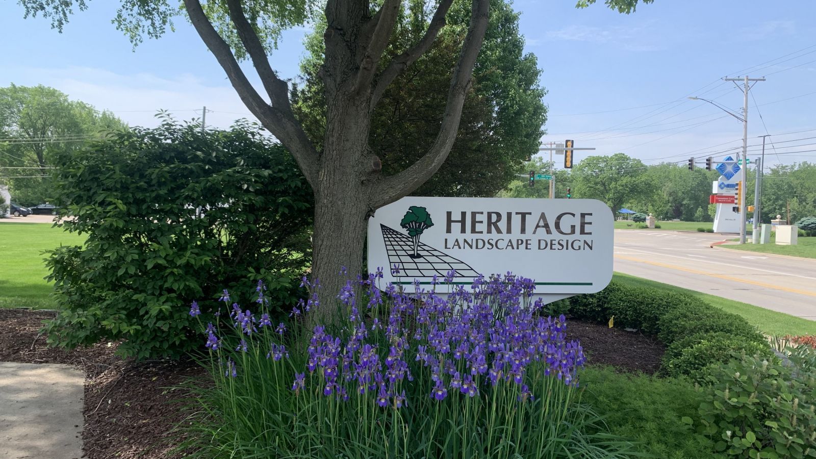 A sign for heritage landscape design is surrounded by purple flowers.