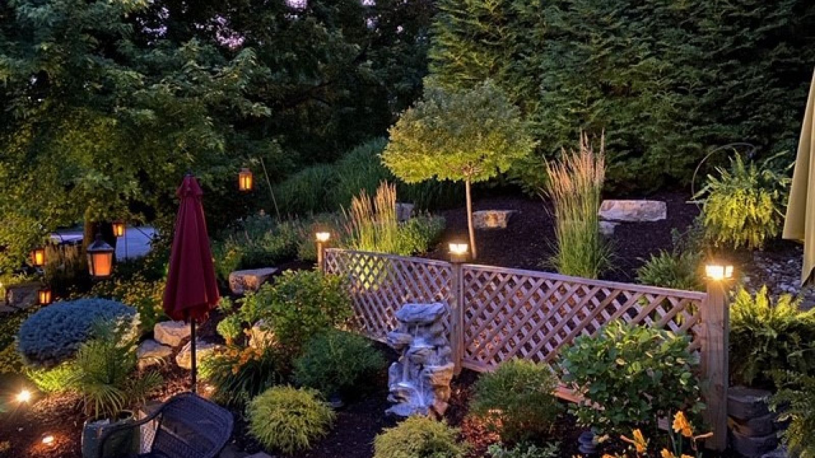A backyard garden illuminated by warm lights at dusk. Features include a decorative lattice fence, lush greenery, and a red umbrella.