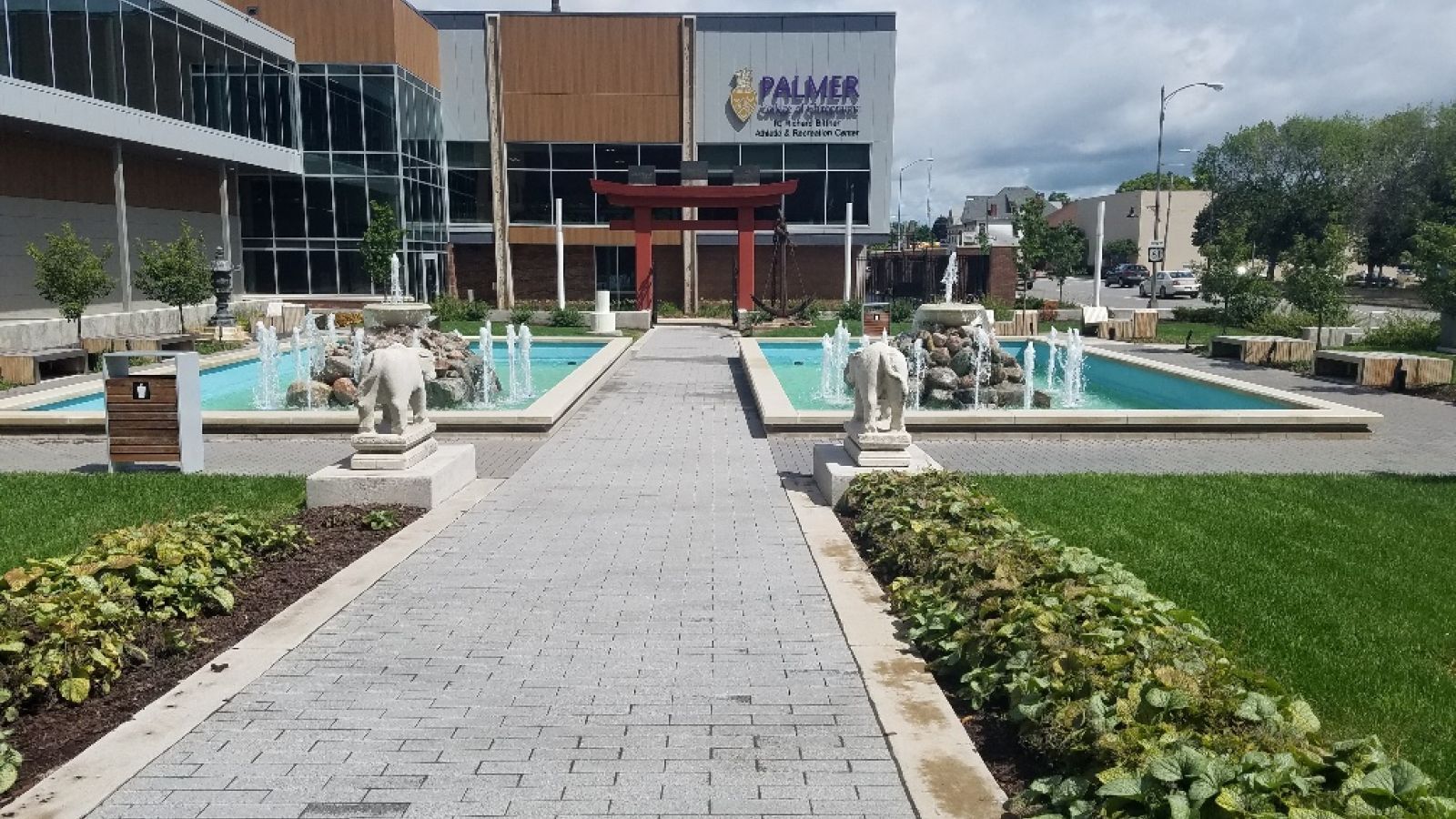 A paved walkway leads to a building, flanked by rectangular water features with statues. The building reads