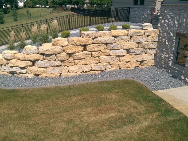 Stone retaining wall with landscaping, grass, and a gravel border next to a house exterior.