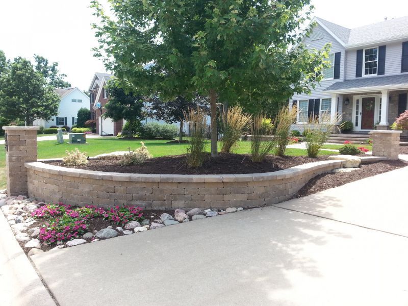 Curved stone retaining wall with a tree and landscaping in front of a house. Pink flowers and rocks line the driveway.