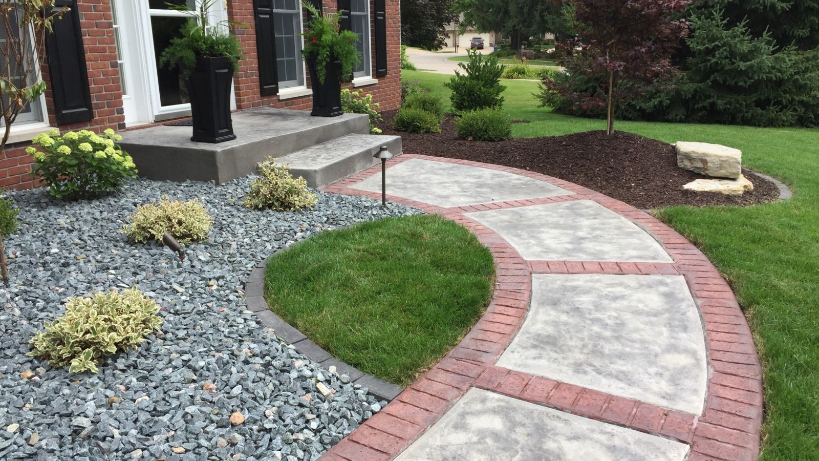 Pathway leading to a house with brick edging, surrounded by green grass, grey gravel, and small bushes.
