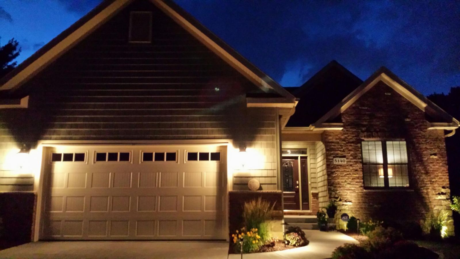 A house lit up at night with garage and walkway lights. Brick and siding exterior, with dark blue night sky.